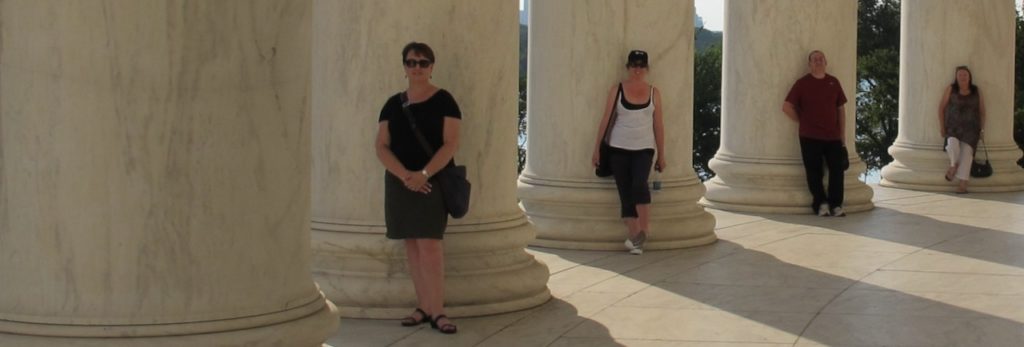 Image is of four people leaning against large ornate pillars.  They are shaded and the sun streams through the gaps of the pillars.