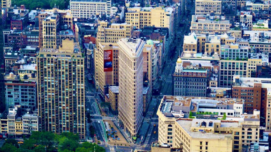 Image of the Flatiron Building in NYC from above and a distance away.  The flatiron thin edge faces forward and two streets fan out.  Other buildings fill the frame.behind it.