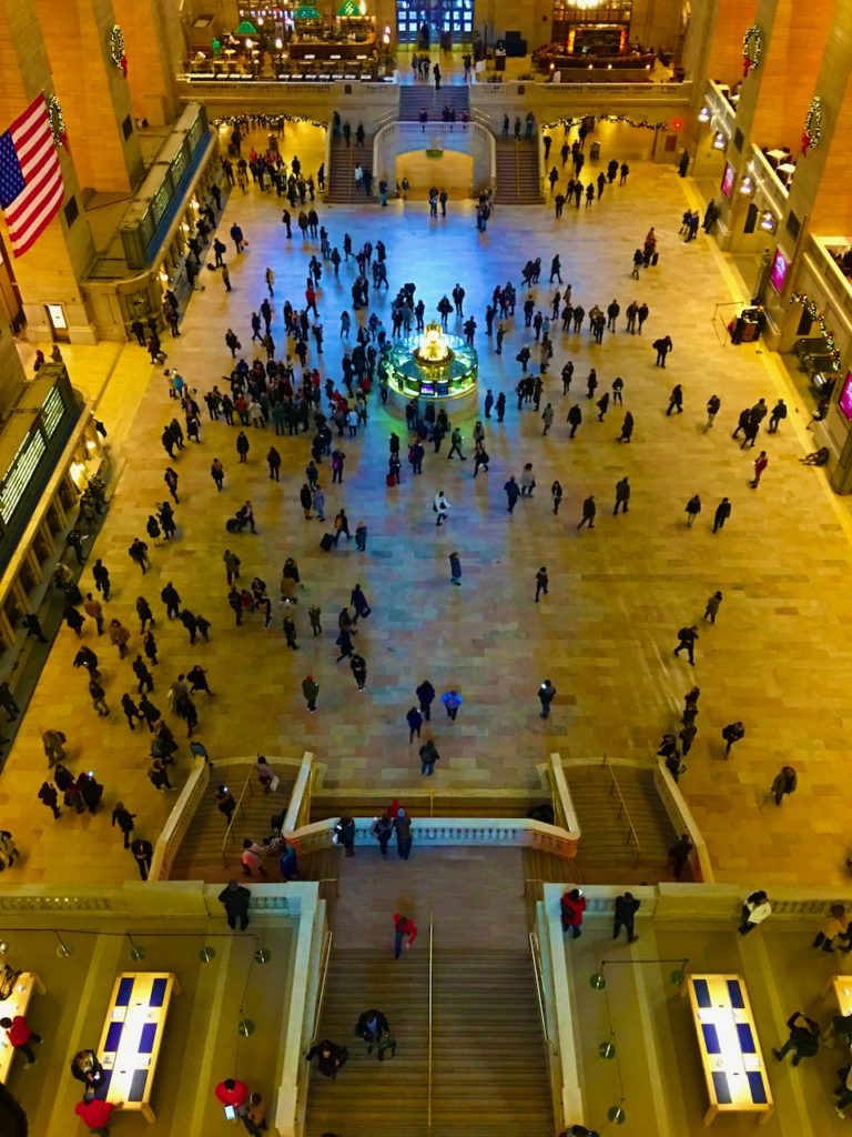Image form internally above Grand Central.  Showing people going about their business on the ground. The vast windows at the far end cast a blue glow on the ground