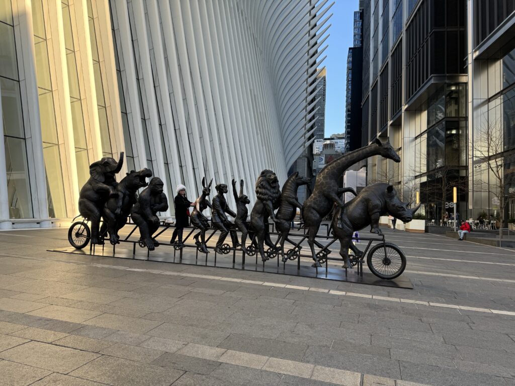 Image of sculpted. Animals on a bike in bronze. Elephant leads, followed by giraffe, zebra, lion, monkey, dog, rabbit, empty seat (human sitting there in photo), ape, hippo, elephant