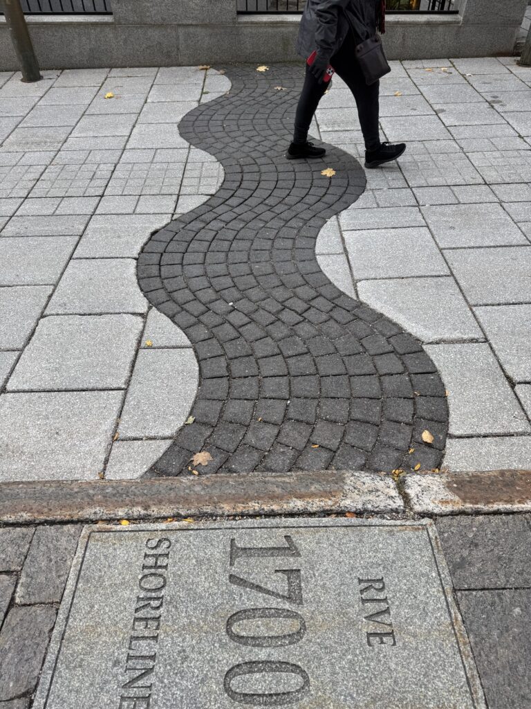 A cobbled toad showing the numbers 1700. The road has a wavy line of black stones surrounded by white. The wave represents the river bank in 1700.