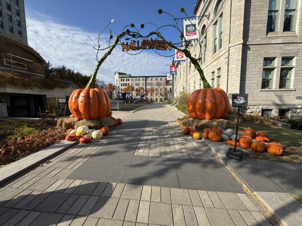 Two massive pumpkins. Each are 6 feet tall. A sign between them says Halloween. The garden behind is ready for a party