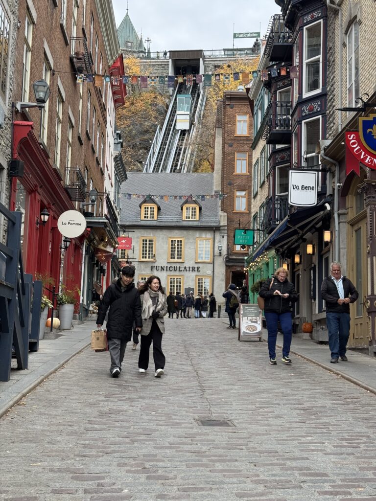 A quaint street in Quebec City. Each side of the street has colourful store fronts. There are people walking up and down the street. In the distance a building sits in front of a steep hill up the you can see a vernacular. A type of train that takes people up the hill in stead of steps.