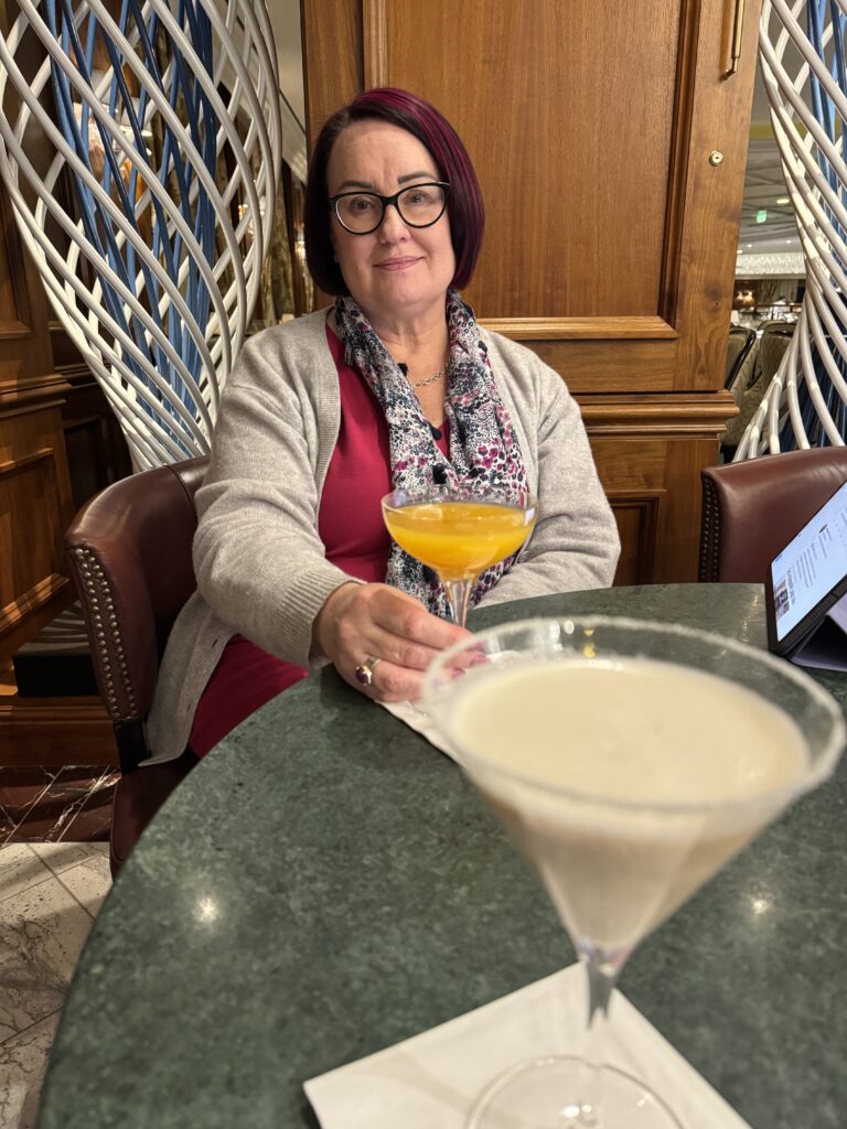 Mardi sitting enjoying a cocktail. She wears a grey cardigan over a pink dress with a grey and white scarf. Her drink is in a martini glass and orange in colour.
