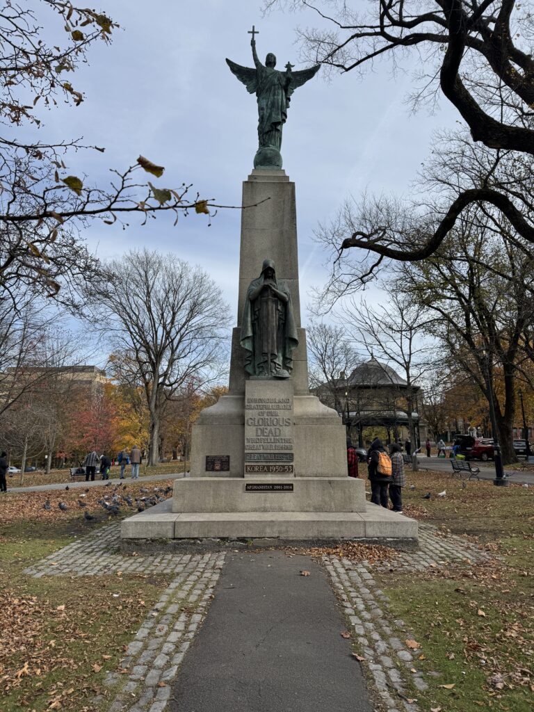 A statue honoring war veterans. A large female figure stophold a cross aloft. It’s about 5 metres tall and The statue is on a concrete plant a shrouded figure below bows his head. Part of the inscription reads glorious dead there are people standing at the base smoking marijuana