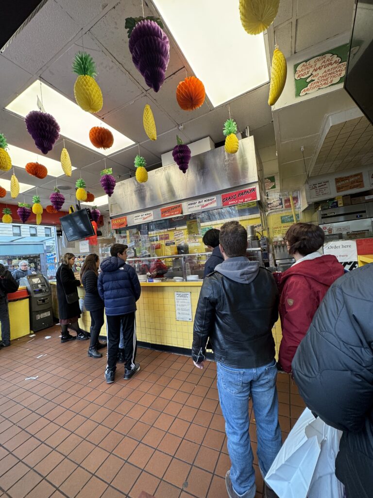 People in gray’s papaya queuing for hot dogs. Fruit, fake, hangs from the ceiling 