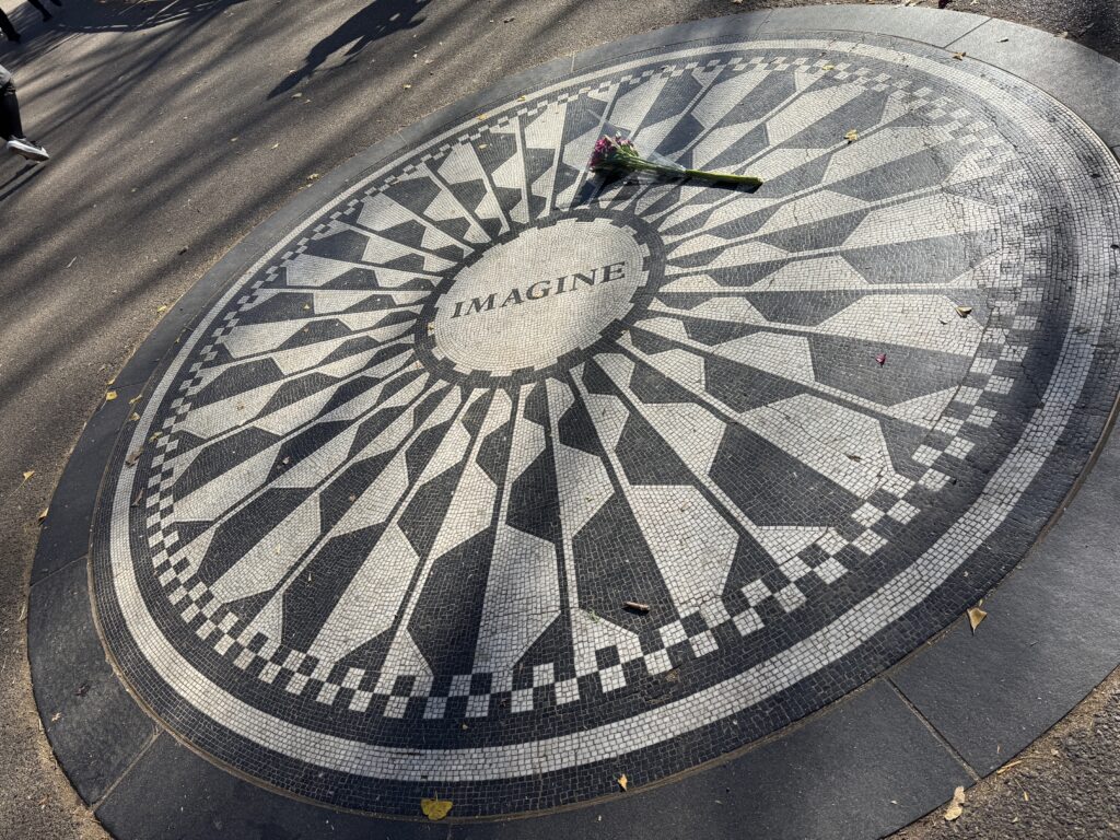 The imagine memorial. Tiles on the ground in a circular pattern, the word imagine in the centre ca single rose, real, lies on the ground above imagine.