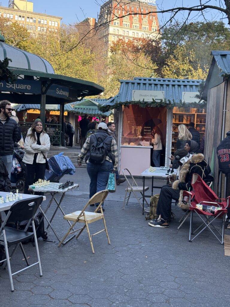 People sitting at tables in the markets drinking and eating. Several stalls can be seen with hoods on show. 
