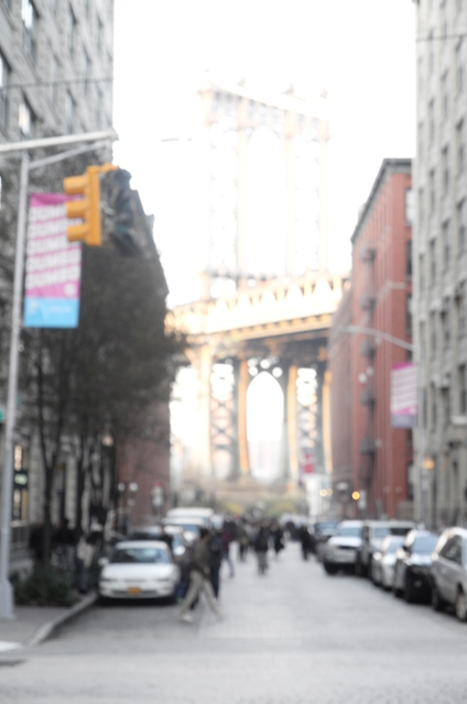 An image from Brooklyn, looking between two warehouses, which frame the Manhattan bridge. The image is washed out and blurry. It is a bright sunny day and the sky is a deep blue. Image was taken on November 23, 2019 at around 3.00pm